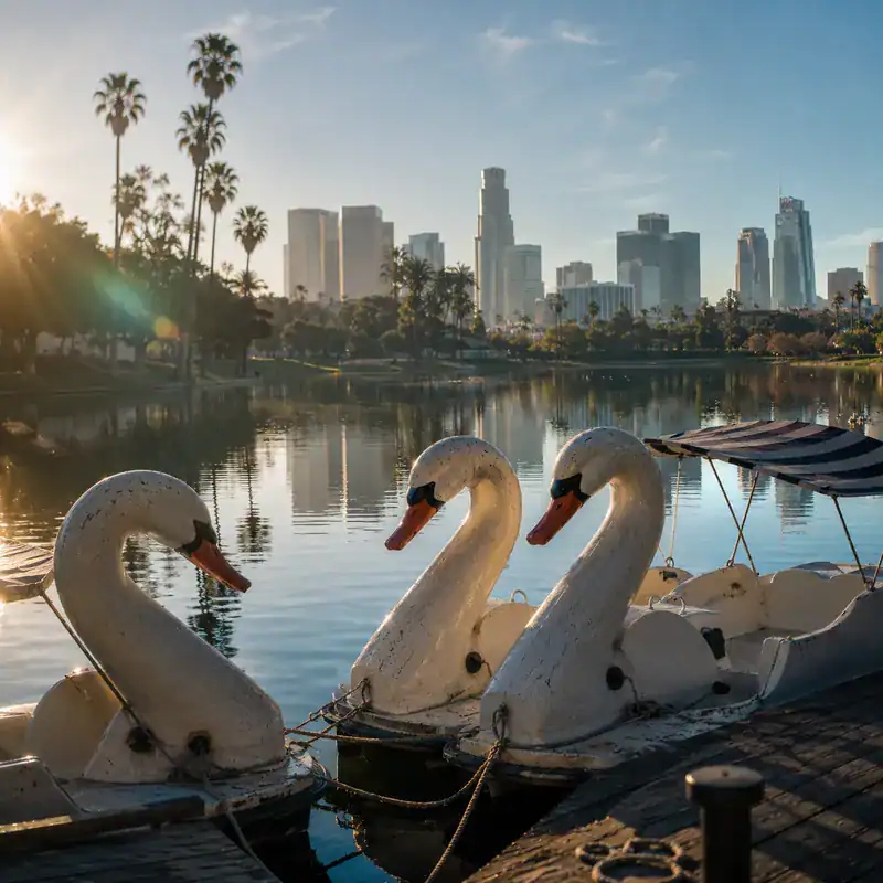 Swan boats on a lake with Los Angeles skyline in the background at sunset.